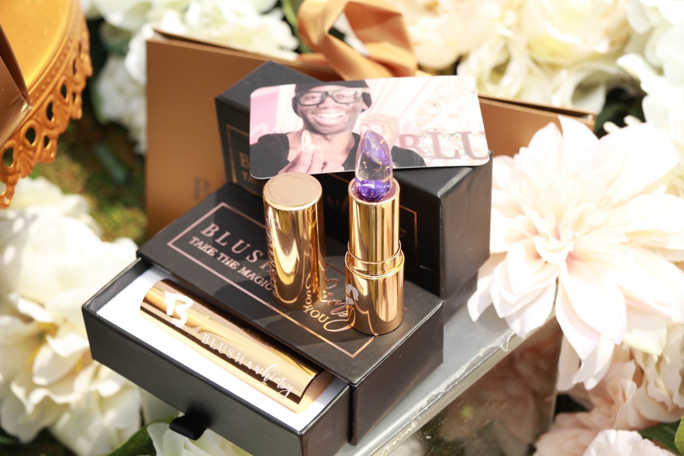 A purple flower lipstick displayed with branded boxes and white flowers at a Blush & Whimsy event