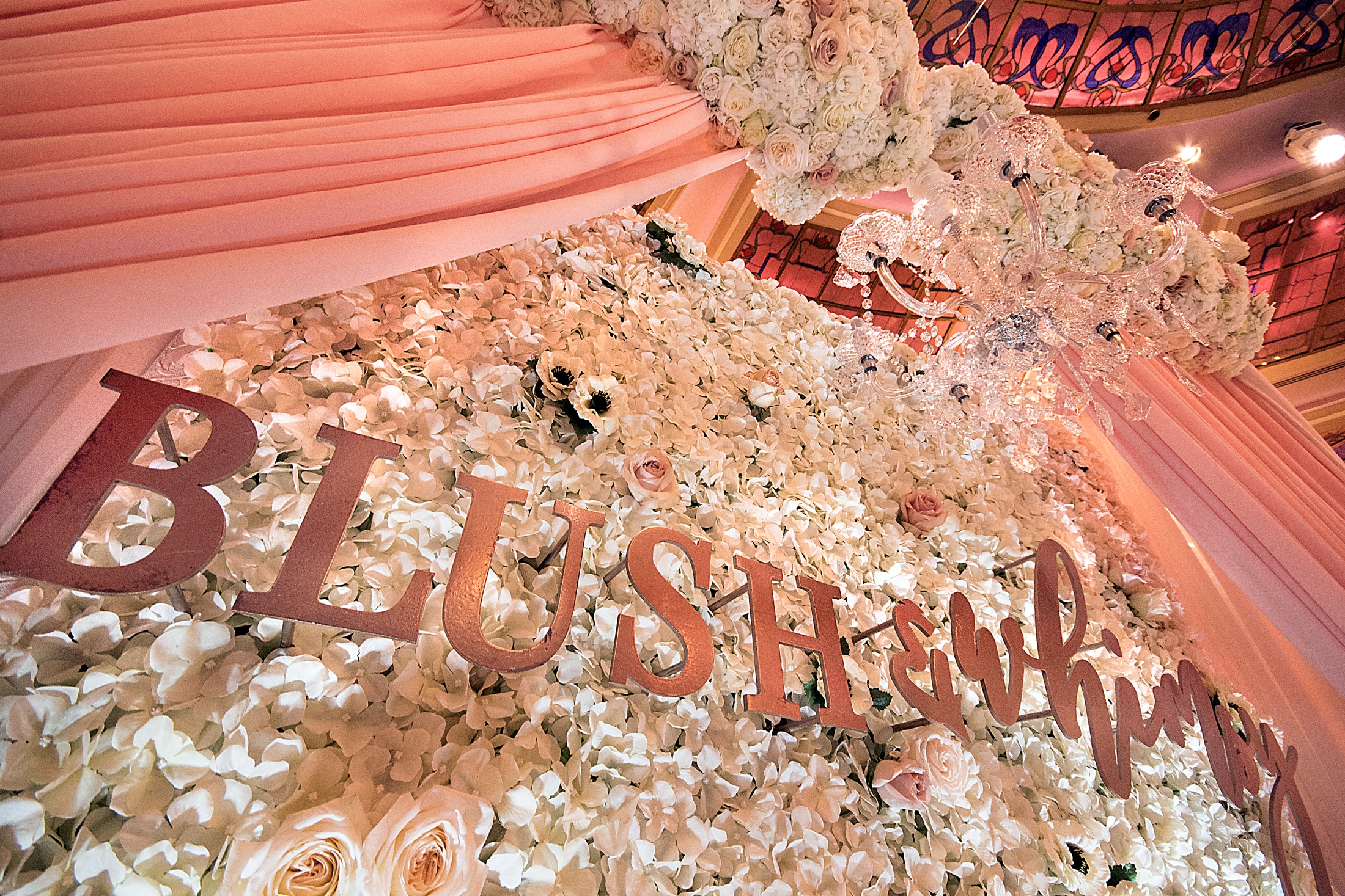 Rose-gold Blush & Whimsy lettering on a flower-covered event wall beneath a glass chandelier